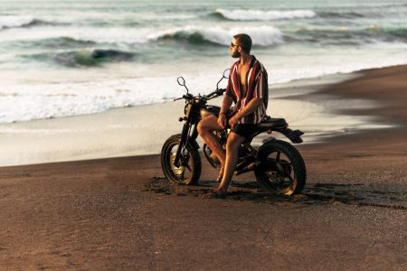 An attractive lonely man with a bike on the beach meets the sunset. Stylish man with a bike by the sea. Travel on a bike. Travel to Bali. A custom bike on the tropical beach.